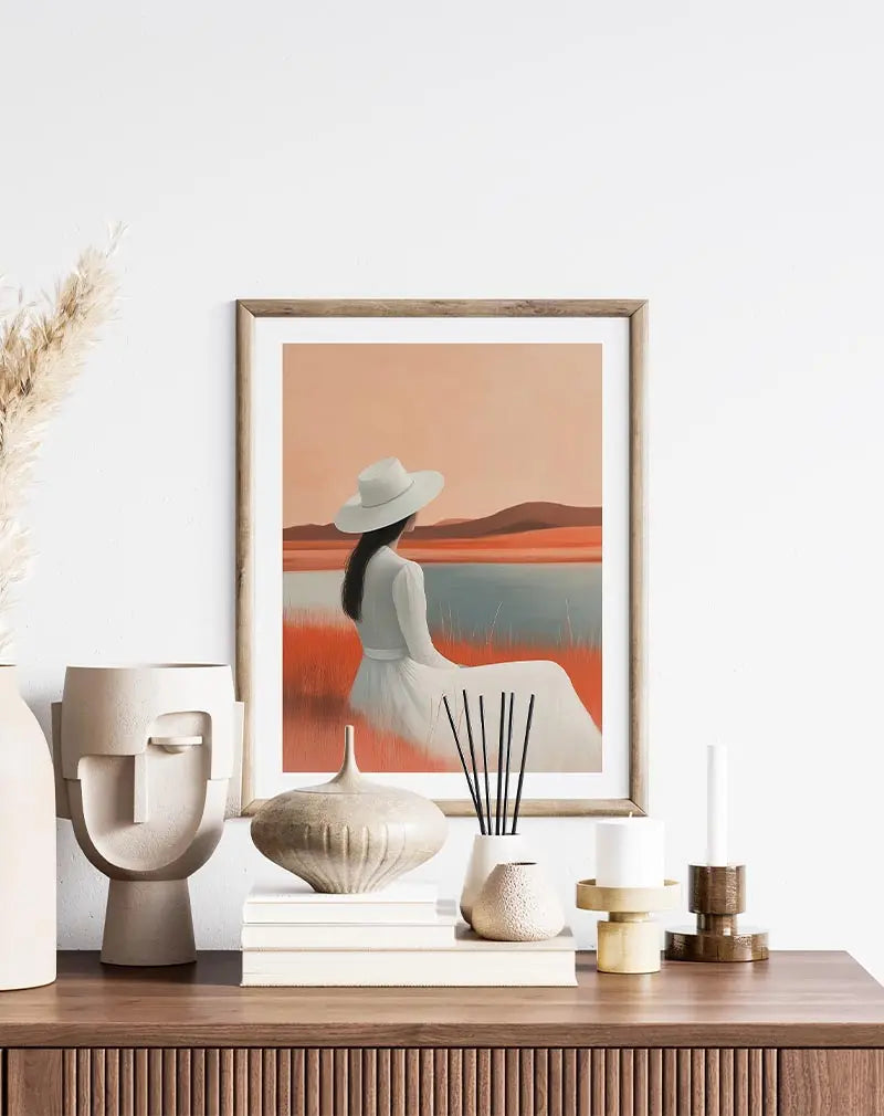 A serene illustration of a woman in a white dress and hat sitting by a calm lakeside with soft orange and brown hues. Perfect wall art for creating a peaceful, reflective atmosphere in any space